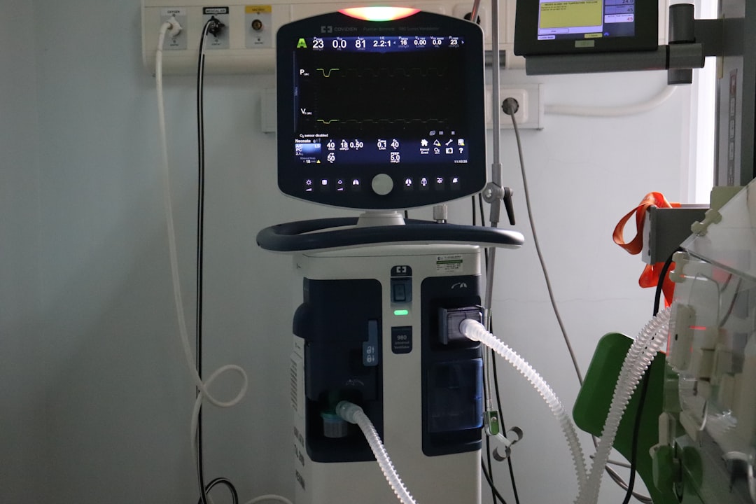 A clinician works late at a dim hospital workstation while an unattended monitor glows in an empty adjacent room