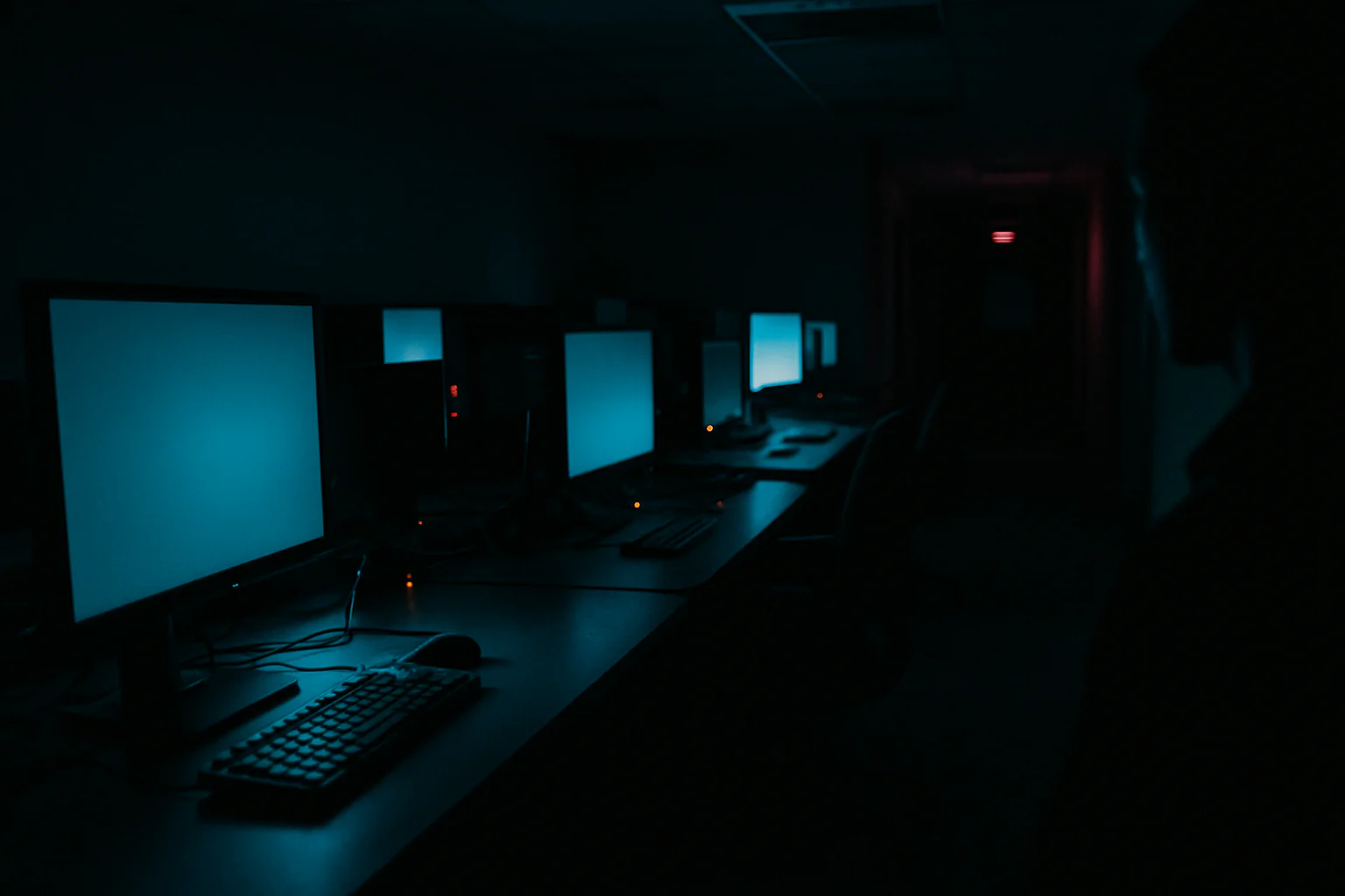 A dim after-hours telehealth operations room lit by monitor glow and indicator LEDs, with a silhouetted figure near the edge of the frame.