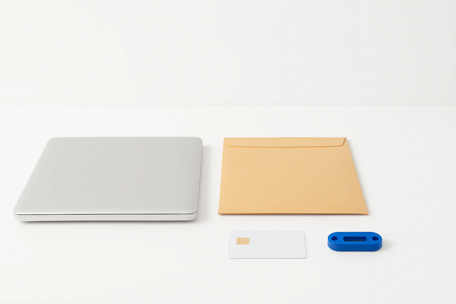 A bright studio still life shows a laptop, file folder, smart card, and blue security token arranged on a white seamless surface.